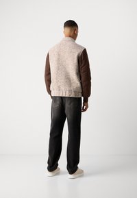 Uomo che indossa una giacca con maniche trapuntate marroni e schiena di tessuto crema, jeans neri e sneakers beige, in piedi di fronte a un muro bianco.