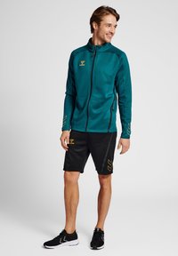 Tealte ritsjack met zwarte accenten en geel logo, gecombineerd met zwarte shorts met een geel chevronpatroon en zwarte sportschoenen.