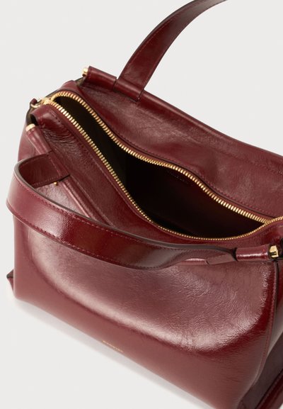 Sac à main en cuir rouge foncé avec une fermeture éclair dorée partiellement ouverte et une large bandoulière sur un fond clair uni.