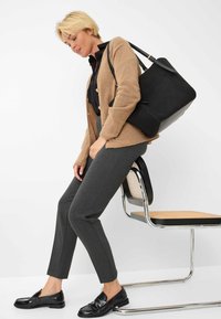 Borsa a tracolla in pelle nera, cardigan beige in maglia con bottoni, camicia nera, pantaloni grigi su misura, mocassini neri lucidi, seduto su una sedia con seduta intrecciata.