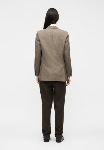 Γυναίκα με μακριά σκούρα μαλλιά φοράει καφέ υφασμάτινο σακάκι, σκούρα παντελόνια και μαύρα παπούτσια, στέκεται με την πλάτη σε απλό φόντο.