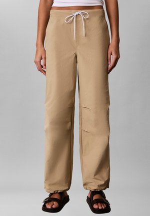 Personne portant un pantalon beige à cordon avec des poignets élastiques et des sandales noires, debout devant un fond gris uni.