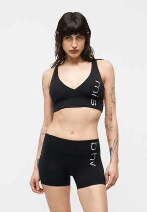 Femme aux cheveux foncés portant un soutien-gorge de sport noir et un short avec un texte de marque blanc, debout devant un fond clair uni.