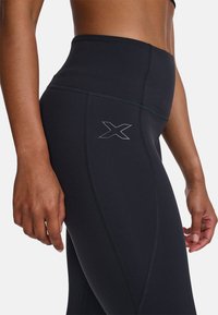 Hochtaillierte schwarze Leggings aus dehnbarem Material, mit einem silbernen X-Logo und kontrastierenden Nähten für zusätzliche Details und Definition.