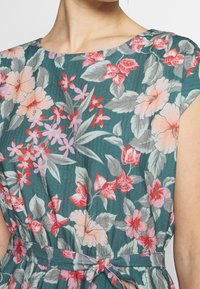 Abito floreale con sfondo teal, caratterizzato da grandi fiori rosa e rossi e foglie verdi; include un dettaglio in vita con cravatta e maniche a pipistrello.