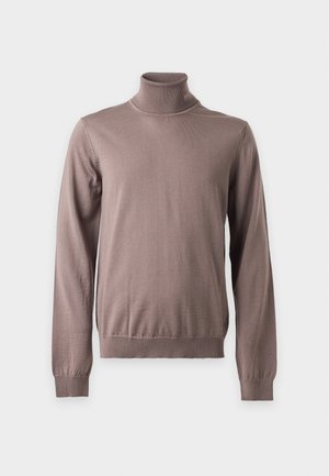 Homme debout, les mains dans les poches, portant un pull à col roulé marron, un pantalon marron foncé et des mocassins noirs et beiges.