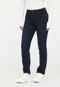 Vaqueros ajustados en denim azul oscuro con una cintura alta. Presentan costuras rojas y bolsillos frontales. Combinados con zapatillas blancas.