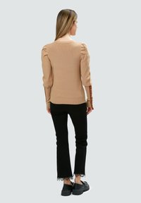 Beige gestrickter Pullover mit puffigen Ärmeln, Rundhalsauschnitt und braunen Akzenten an den Bündchen; kombiniert mit schwarzen, ausgestellten Jeans mit Spitzenbesatz.