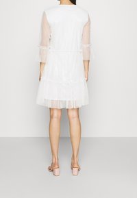 Robe blanche avec manches longues transparentes et accents en tulle à volants. Présente un col rond et arrive au-dessus du genou. Portée avec des sandales à talons blocs roses.