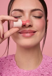 Botella aplicadora de rubor sostenida cerca de la nariz, con un producto cremoso color coral rosado aplicado en la piel. Contraste entre la tapa texturizada rosa y la piel suave.