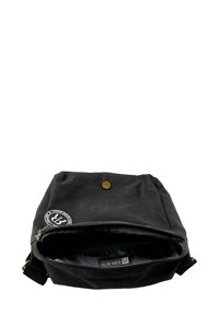 Black canvas bag with a top flap, brass snap closure, and a zippered compartment. Interior features a patterned lining and a label.