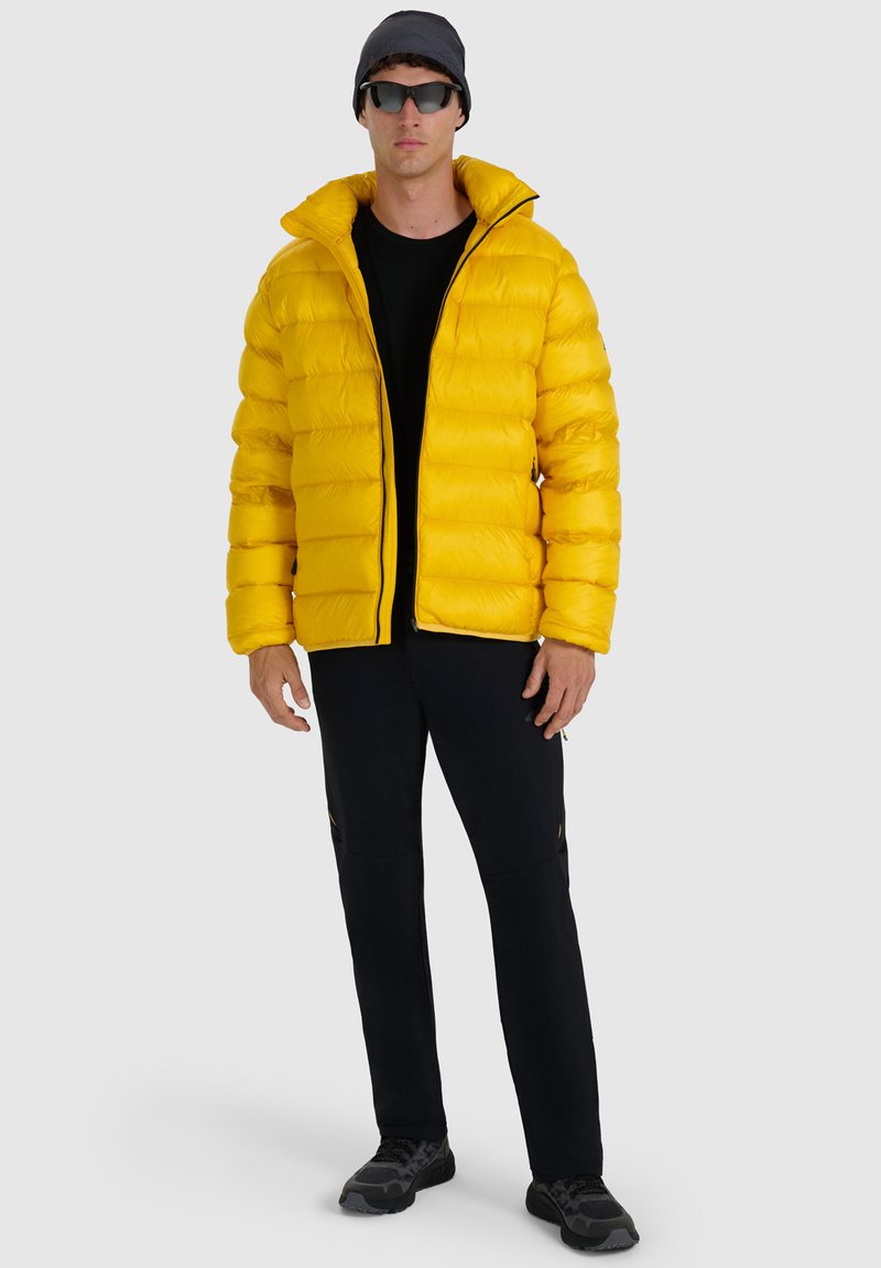 Homme debout face à l'avant portant une doudoune jaune, un pantalon noir, un t-shirt noir, des lunettes de soleil, un bonnet et des baskets noires sur un fond clair.
