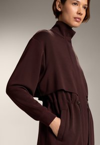 Bordó, cipzáros overál, puha anyagból készült, magas gallérral, rugalmas derékkal és oldalsó zsebekkel. Széles ujjak jellemzik, valamint réteges kialakítással rendelkezik.