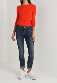 Camiseta roja de manga larga con textura acanalada combinada con jeans de mezclilla azul oscuro. Los jeans tienen un corte ajustado y están enrollados en los puños, complementados con zapatillas blancas.