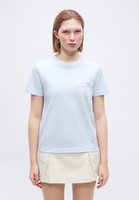 Γυναίκα με κοντά μαλλιά που φοράει ένα γαλάζιο t-shirt Calvin Klein και μια μπεζ πλισέ μίνι φούστα, στέκεται μπροστά σε λευκό φόντο.