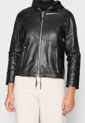 Personne portant une veste en cuir noire zippée avec une capuche et des poches zippées, associée à un pantalon beige clair.