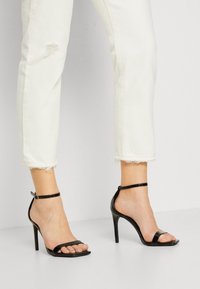 Sandalias de cielo alto de charol negro con dos correas anchas y una correa en el tobillo, sobre unos jeans cortos desgastados blancos.