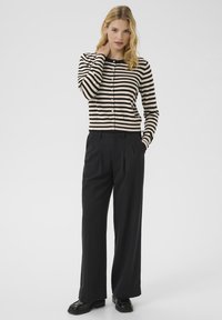 Cardigan rayé noir et beige à manches longues et petits boutons, associé à un pantalon large noir taille haute et des chaussures noires.