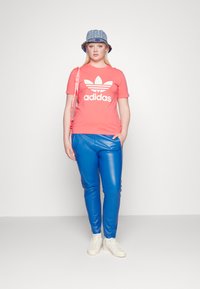 Korallis tshirts ar baltu Adidas logo, spīdīgas zilo mākslīgās ādas bikses, baltas kedas un džinsu raksta kausa cepure.