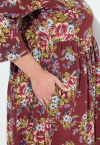 Vestido floral en burdeos con mangas cortas. Presenta flores coloridas en rosa, azul y verde. Detalle de bolsillo en el lado izquierdo.