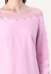 Top de manga larga en tela de color rosa claro con un yugo transparente y bordes ondulados en las mangas. Textura suave, cuello redondo simple.