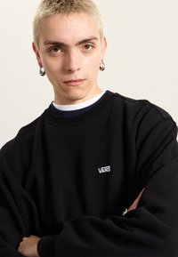 Black sweatshirt with rounded neckline and white Vans logo. Model has short blonde hair, silver hoop earrings, and arms crossed.