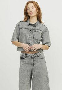 Chaqueta corta de mezclilla gris con un collar estructurado, cierre de botones, bolsillo frontal y mangas cortas, combinada con pantalones de mezclilla gris de cintura alta.