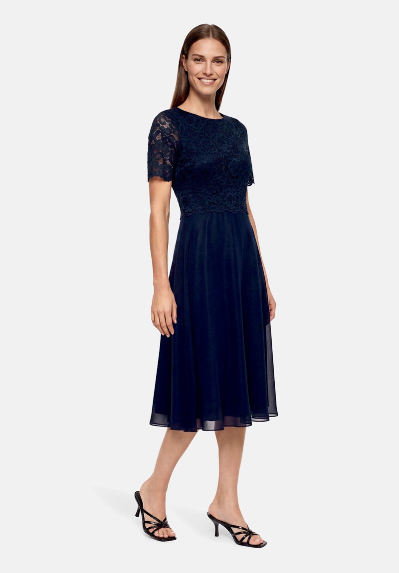 Donna che indossa un vestito blu navy al ginocchio con maniche corte in pizzo e corpetto aderente, abbinato a sandali neri con tacco.