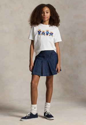 Dziewczyna z kręconymi włosami ubrana w białą koszulkę z nadrukiem misia, plisowaną ciemnoniebieską spódnicę z denimu, białe skarpetki i granatowe trampki, stoi.