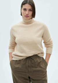 Beige højhalset sweater lavet af blødt materiale med ribbede manchetter og halsudskæring. Kombinationen med mørkegrønne trækbåndsbukser.