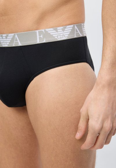 Άνδρας που φοράει μαύρο εσώρουχο με γκρι ζώνη μέσης με το λογότυπο Emporio Armani, εμφανίζοντας τον κορμό, το άνω μέρος του μηρού και το χέρι χαλαρό στο πλάι.