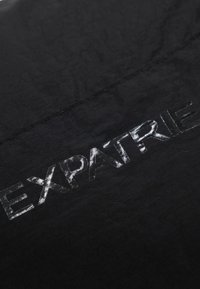 Sort stof prydes af det prægede tekst "EXPATRIÉ" i en blank finish, hvilket skaber en subtil kontrast mod den teksturerede baggrund.