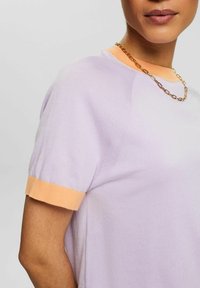 Jasnofioletowa, dzianinowa koszulka z krótkim rękawem i pomarańczowymi ściągaczami. Posiada gładką teksturę oraz okrągły dekolt z delikatnymi przeszyciami.