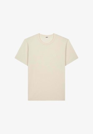 T-shirt beige à manches courtes et col rond avec un petit logo circulaire sur la poitrine gauche et une étiquette JOTT à l'intérieur du col.
