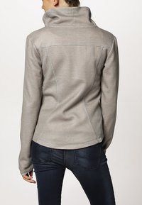 Giacca grigia chiara con una finitura testurizzata, colletto alto e cuciture in contrasto. Design aderente con maniche lunghe e tessuto liscio.