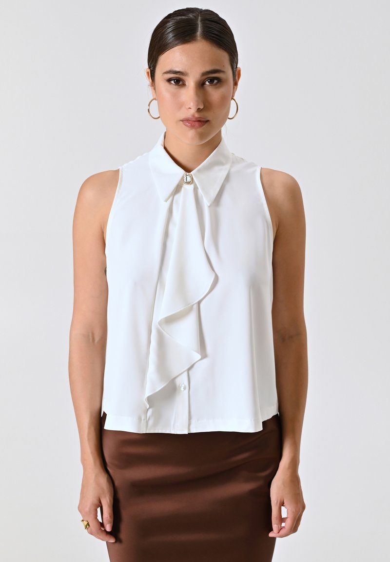 Weiße ärmellose Bluse mit gerafftem Vorderteil, Knopfverschluss und Kragen. Glattes Gewebe, ergänzt durch einen dekorativen Edelstein am Hals.