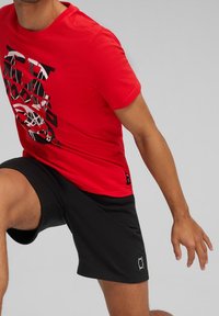 Červené tričko s černým grafickým textovým designem, v kombinaci s černými šortkami. Tkanina vypadá hladce, má standardní střih a kulatý výstřih.