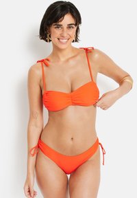 Ryškiai oranžinė bikini kostiumėlis, kuriame yra raukinėtas bandeau tipo viršus su reguliuojamomis petnešėlėmis ir atitinkamos kelnaitės su raištelių detalėmis šonuose. Lygi, tampra medžiaga.