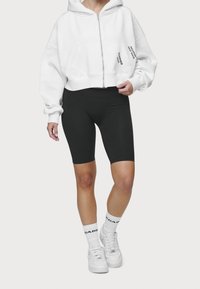 Sudadera blanca corta con cremallera frontal y dos bolsillos, combinada con pantalones cortos ajustados negros y zapatillas blancas con acentos negros.