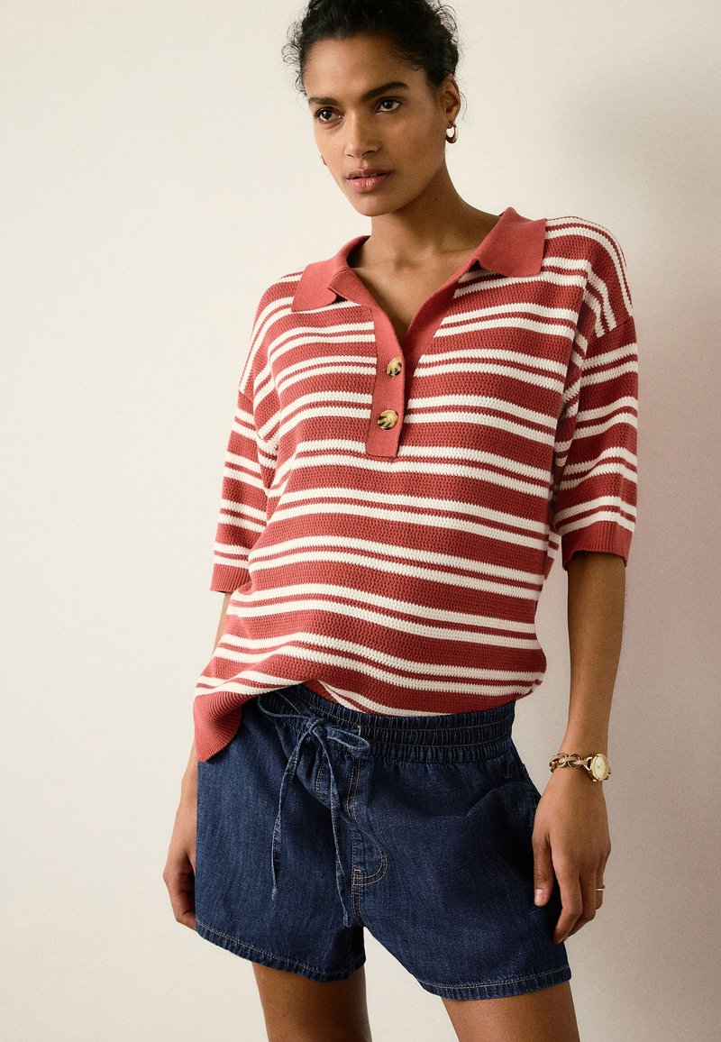 Mujer que lleva un top de punto de manga corta a rayas rojas y blancas con botones y pantalones cortos de mezclilla azul oscuro con cordón.