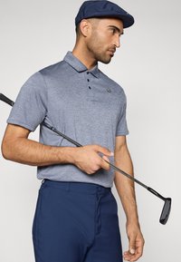 Μπλε πόλο πουκάμισο με γιακά, τρία κουμπιά και λογότυπο. Συνδυασμένο με ναυτικό παντελόνι γκολφ. Κρατάει ένα μπαστούνι γκολφ και φοράει ένα ναυτικό καπέλο.