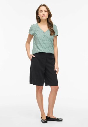 Sieviete stāv ar vienu roku kabatā, valkājot gaiši zaļu īss piedurkņu blūzi ar rakstu, melnas pusceļa garuma šortus un melnus dzīvokļveida apavus.