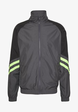 Giacca a vento nera e grigia con un colletto alto, chiusura con zip, polsini elastici e due strisce verdi sulle maniche. Design leggero e sportivo.