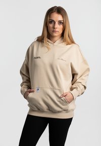 Beige Kapuzenpullover mit großer Vordertasche, Kordelzug und gerippten Bündchen. Verfügt über dezentes Branding auf der Brust und ein Stoffetikett.