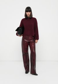 Burgunderfarbener Strickpullover mit schwarzem Kragen, kombiniert mit glänzenden roten Hosen im Krokodilmuster. Hält eine schwarze Handtasche und trägt schwarze Schuhe.