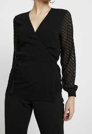 Vrouw die een zwarte wikkelblouse draagt met doorschijnende, bedrukte lange mouwen en een zwarte broek, staand tegen een effen lichte achtergrond.