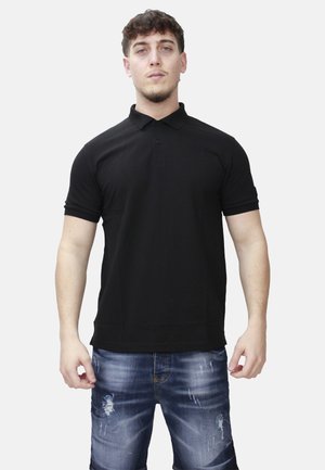 Uomo in piedi che indossa una polo nera a maniche corte e pantaloncini di jeans blu slavati contro uno sfondo chiaro e semplice.