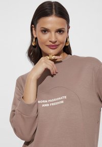 Brązowa bluzka z dołem w kształcie łuku, z białym napisem "URODZONY Z PASJĄ I WOLNOŚCIĄ" na przodzie. Miękka tkanina, okrągły dekolt, długie rękawy.