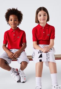 Två barn sitter på en bänk, iförda röda Lacoste-pikétröjor. Den ena har gråa shorts, och den andra har en mönstrad plisserad kjol och vita sneakers.