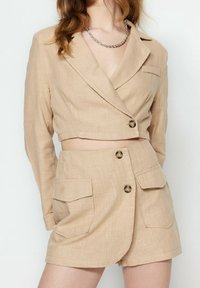Giacca cropped beige con maniche lunghe e colletto a bavero, abbinata a pantaloni corti a vita alta. Entrambi presentano tasche con pattina e dettagli con bottoni.
